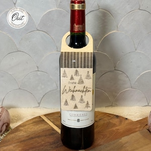May include: A wooden wine bottle tag with a laser-cut design of Christmas trees and the text "Frohe Weihnachten".