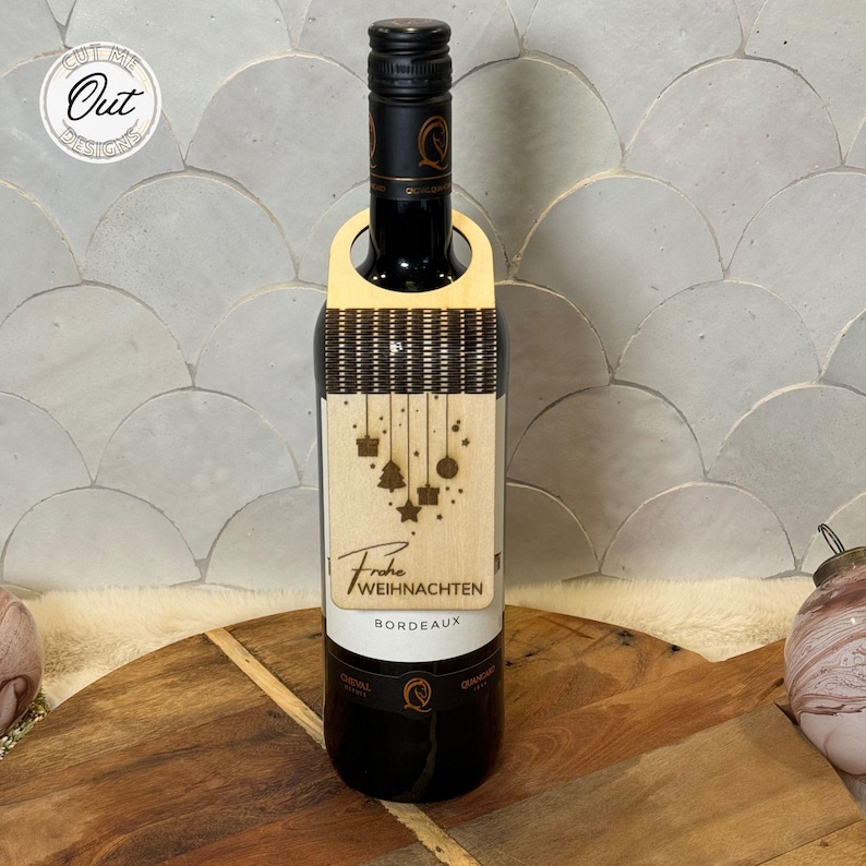 May include: A wooden wine bottle tag with a Christmas theme. The tag features a hanging ornament design with a Christmas tree, stars, and presents. The text "Frohe Weihnachten" is printed on the tag, along with the word "Bordeaux".