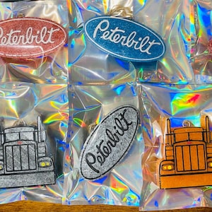 May include: Eight Peterbilt truck themed keychains in various colors and finishes. The keychains feature a Peterbilt logo and a miniature truck design. The keychains are packaged in clear plastic bags.