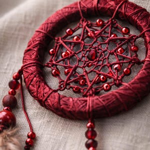 May include: A handcrafted, burgundy dreamcatcher. The circular frame is wrapped in matching thread, with a web of burgundy string and red beads. Decorative beads and feathers hang from the bottom.