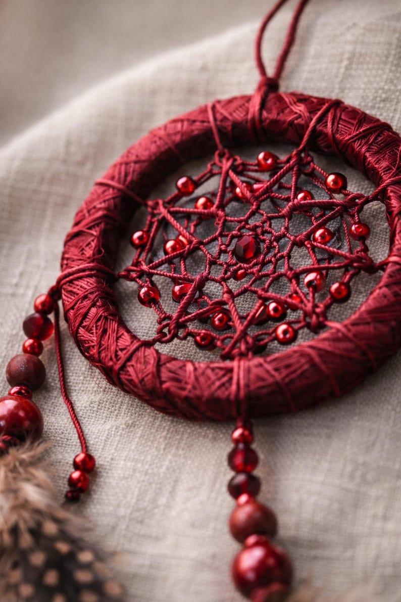 May include: A handcrafted, burgundy dreamcatcher. The circular frame is wrapped in matching thread, with a web of burgundy string and red beads. Decorative beads and feathers hang from the bottom.