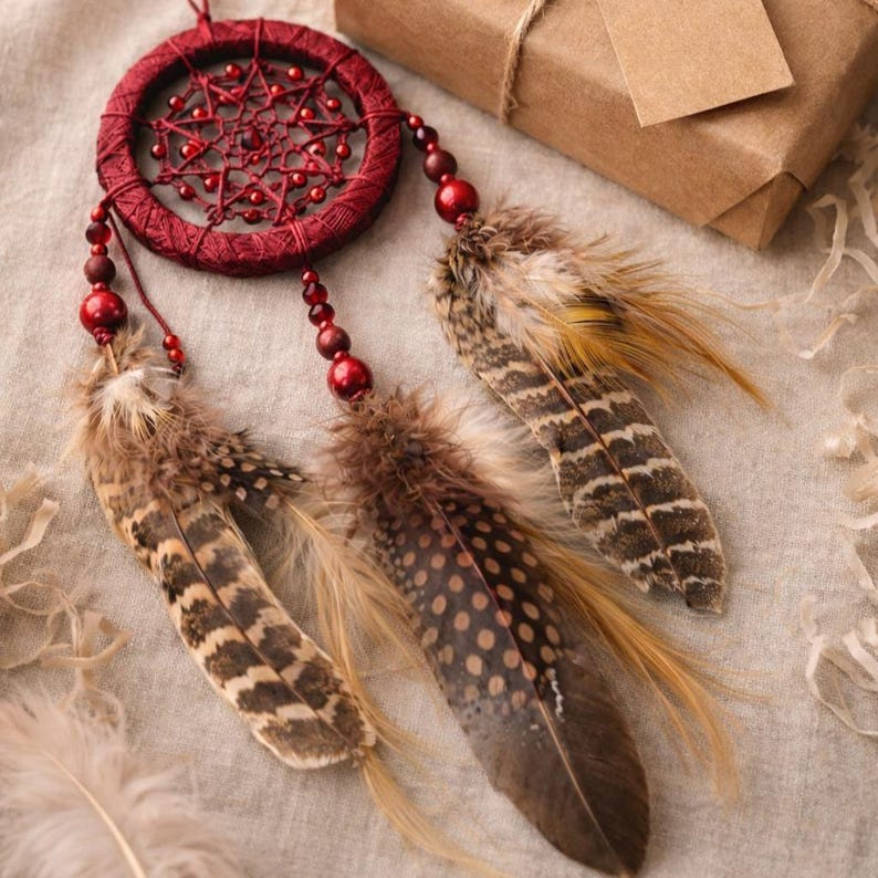 May include: A handcrafted red dreamcatcher with a circular woven frame and beaded accents. Brown and tan feathers hang from the bottom. A gift box with a blank tag is in the background.