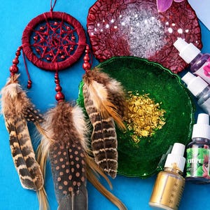 May include: A burgundy dreamcatcher with brown and white feathers hangs near a green dish with gold flakes. A red, textured resin coaster and several bottles of liquid are also present. The background is a bright blue.