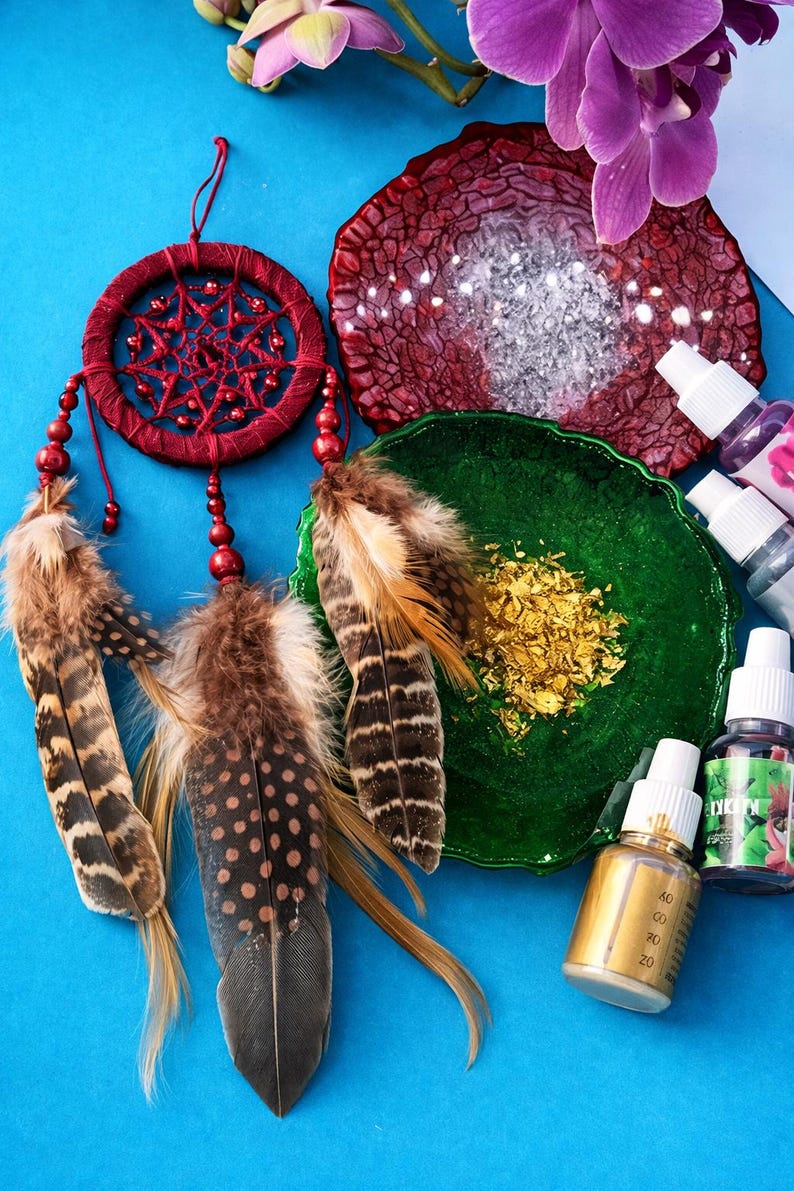 May include: A burgundy dreamcatcher with brown and white feathers hangs near a green dish with gold flakes. A red, textured resin coaster and several bottles of liquid are also present. The background is a bright blue.