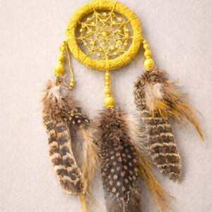 May include: A handcrafted dreamcatcher with a yellow woven hoop, adorned with yellow beads and three brown and tan feathers. The dreamcatcher is suspended by a yellow string, creating a decorative wall hanging.