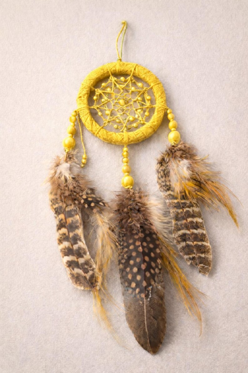 May include: A handcrafted dreamcatcher with a yellow woven hoop, adorned with yellow beads and three brown and tan feathers. The dreamcatcher is suspended by a yellow string, creating a decorative wall hanging.