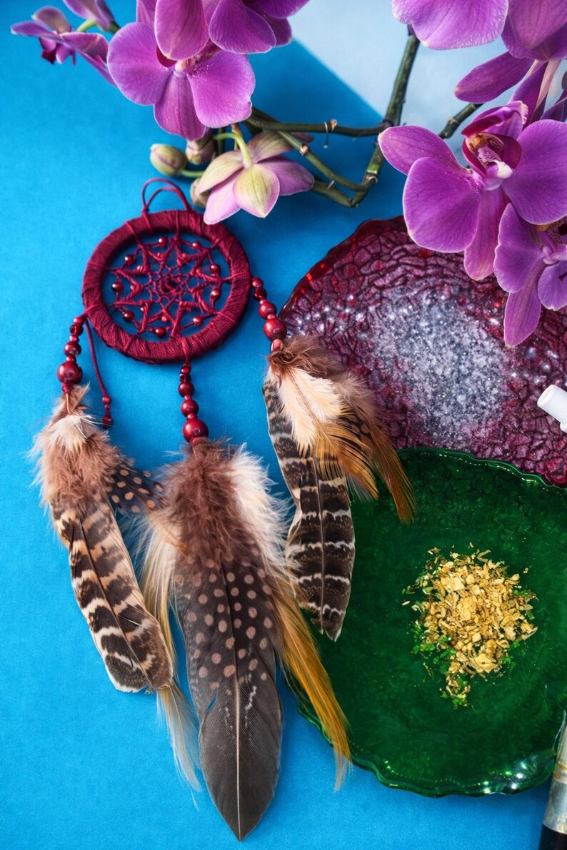 May include: A burgundy dreamcatcher with brown and tan feathers hangs against a blue background. Purple orchid flowers and a green and burgundy decorative plate are also in the image. The dreamcatcher has red beads.