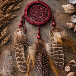 May include: A burgundy dreamcatcher with a woven web design and beaded accents. The dreamcatcher features three decorative feathers with brown and tan patterns. Seashells and dried plants surround the dreamcatcher on a wooden surface.