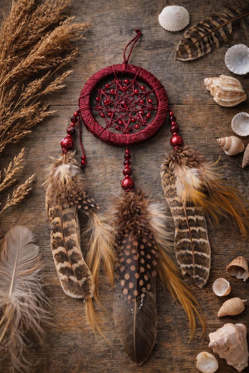 May include: A burgundy dreamcatcher with a woven web design and beaded accents. The dreamcatcher features three decorative feathers with brown and tan patterns. Seashells and dried plants surround the dreamcatcher on a wooden surface.