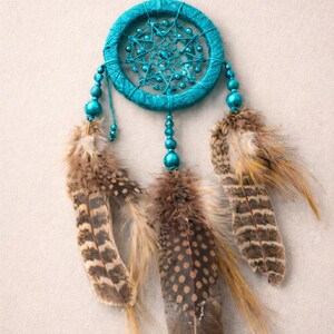 May include: A turquoise dreamcatcher with a woven circular frame and beaded accents. Three brown and tan feathers hang below. The dreamcatcher is suspended against a neutral background.