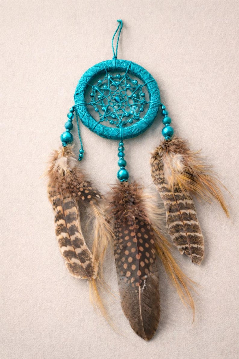 May include: A turquoise dreamcatcher with a woven circular frame and beaded accents. Three brown and tan feathers hang below. The dreamcatcher is suspended against a neutral background.