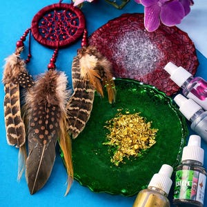 May include: A red dreamcatcher with brown and tan feathers, a green glass plate with gold flakes, and small bottles of liquid are arranged on a blue surface. Purple orchids are in the background.