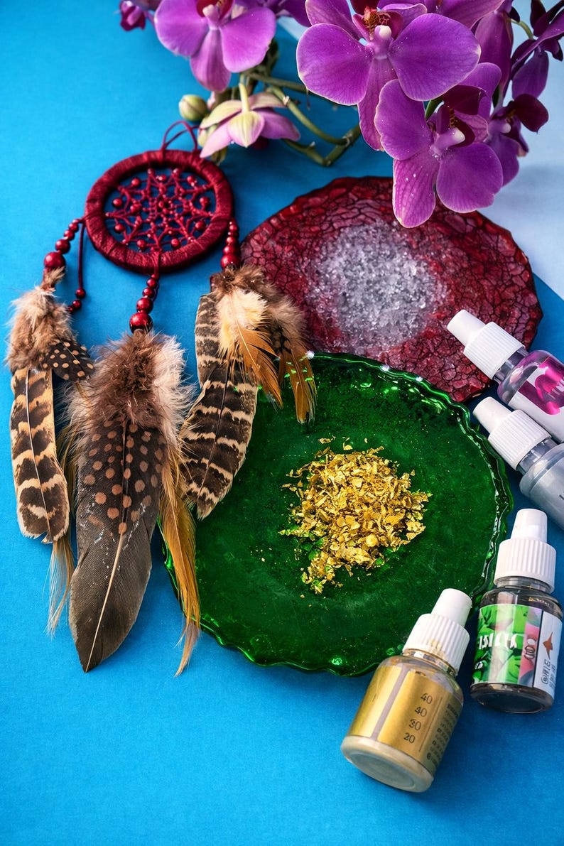 May include: A red dreamcatcher with brown and tan feathers, a green glass plate with gold flakes, and small bottles of liquid are arranged on a blue surface. Purple orchids are in the background.