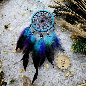 May include: A blue and purple dreamcatcher with black feathers and beads. The dreamcatcher is hanging from a string and is surrounded by white salt crystals. The label "HAND MADE" is visible in the lower right corner of the image.