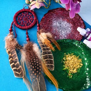 May include: A red dreamcatcher with brown and tan feathers hangs near a purple orchid. A dark red, textured stone and a green dish with gold flakes are also in the image. The background is a bright blue.