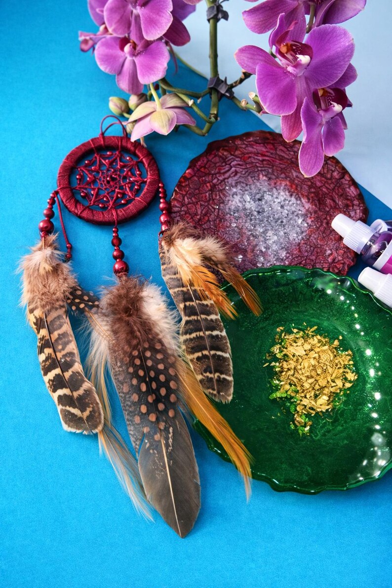 May include: A red dreamcatcher with brown and tan feathers hangs near a purple orchid. A dark red, textured stone and a green dish with gold flakes are also in the image. The background is a bright blue.