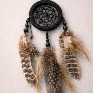 May include: A black dreamcatcher with a woven circular frame and beaded accents. Three feathers in shades of brown and tan hang below. The dreamcatcher is suspended against a neutral background.