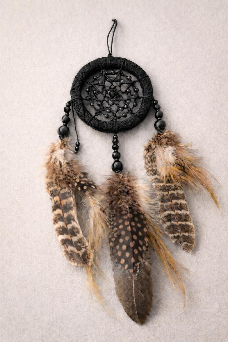 May include: A black dreamcatcher with a woven circular frame and beaded accents. Three feathers in shades of brown and tan hang below. The dreamcatcher is suspended against a neutral background.