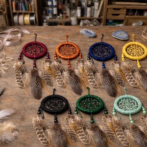 May include: A collection of handmade dream catchers in various colors, including red, orange, blue, yellow, black, green, and turquoise. Each dream catcher features a circular woven web design, adorned with feathers and beads. The dream catchers are displayed on a wooden surface.