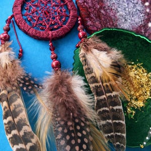 May include: A burgundy dreamcatcher with a woven web design and beaded accents. The dreamcatcher features decorative feathers in shades of brown, tan, and black. The background is a solid blue.