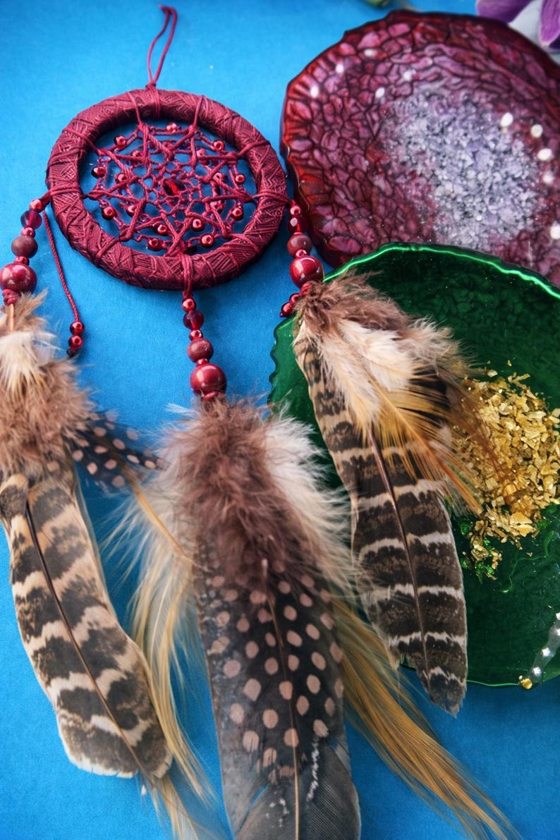 May include: A burgundy dreamcatcher with a woven web design and beaded accents. The dreamcatcher features decorative feathers in shades of brown, tan, and black. The background is a solid blue.