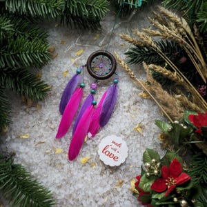 May include: A pink and purple dreamcatcher with feathers and beads hanging from a wooden ring. The dreamcatcher is surrounded by white salt crystals and a small white tag with the text "made with love".