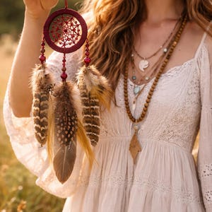 May include: A burgundy dreamcatcher with a web design and hanging feathers. The dreamcatcher is adorned with red beads and brown and tan feathers. The item is held in front of a person wearing a white dress and multiple necklaces.