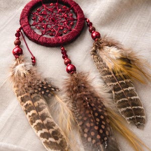 May include: A burgundy dreamcatcher with a woven web design and red beads. Three feathers in shades of brown and tan hang below the circular frame. The dreamcatcher is suspended by a thin red string.