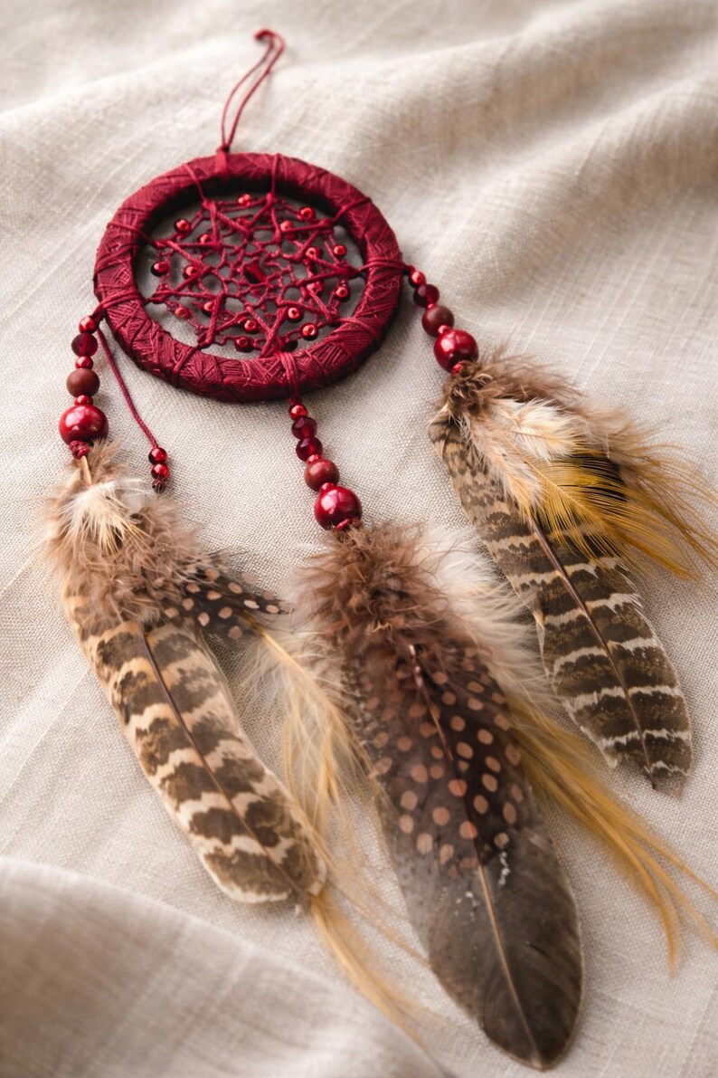 May include: A burgundy dreamcatcher with a woven web design and red beads. Three feathers in shades of brown and tan hang below the circular frame. The dreamcatcher is suspended by a thin red string.