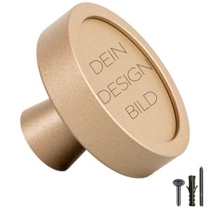 Peut inclure: Un bouton rond de couleur or avec un bord surélevé et le texte "DEIN DESIGN BILD" gravé sur le dessus. Le bouton est fixé à une base plus petite de couleur or.