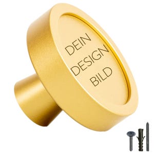Peut inclure: Bouton rond de couleur or avec un bord surélevé et le texte "DEIN DESIGN BILD" gravé sur la surface.