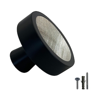 Könnte beinhalten: Schwarzer Schrank-Knopf mit einer runden, silberfarbenen Oberfläche. Der Knopf hat eine gebürstete Metalloberfläche und eine schwarze Basis.