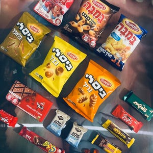 Peut inclure: Une variété de collations dans des sacs et des emballages colorés. Les collations comprennent des chips, des bretzels, des barres chocolatées et des bonbons. Les sacs sont étiquetés en hébreu.