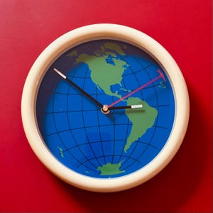 May include: A round wall clock with a cream-colored frame. The clock face features a blue background with a green map of the world. Black and white hands indicate the time, and a red arrow-shaped hand sweeps across the face.