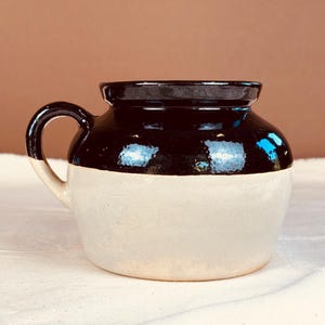 May include: A two-toned ceramic jar with a handle. The top half is a glossy black, while the bottom half is a matte cream color. The jar has a rounded shape and a small handle on the side. It sits on a white surface.