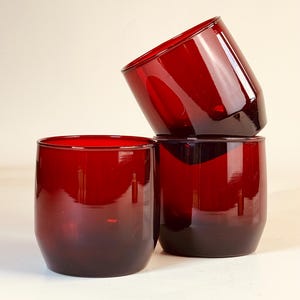 May include: Three deep red glass tumblers with a smooth, cylindrical shape and slightly tapered bases. One tumbler stands upright in the foreground, while two others are stacked vertically behind it, creating a vibrant, translucent display.