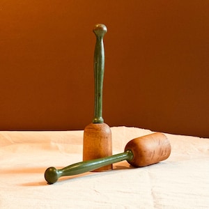 May include: Two wooden mallets with cylindrical heads and long, slender green handles. One mallet stands upright on its flat base, while the other rests horizontally. The wooden heads feature a natural light brown finish with visible grain.