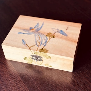 May include: A light-colored wooden box with a hinged lid. The box is decorated with a blue floral design and a metal clasp. The box is sitting on a dark brown wooden surface.