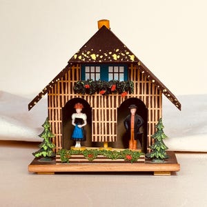 May include: A wooden decorative house with a brown roof and a blue window. The house features two figurines, a woman in a blue skirt and a man in a black coat. Green and red floral accents and two small green trees decorate the house.