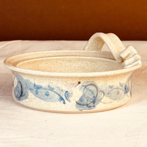 May include: A round, speckled ceramic dish with a single arched handle on one side. The exterior features a hand-painted blue floral vine pattern with dark gray accents. The clay has a light beige, speckled finish throughout the entire piece.
