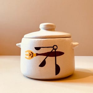 May include: A cream-colored ceramic pot with a lid and two small handles. The pot features a whimsical design of a yellow fork, brown spoon, and black ladle. The pot is likely for kitchen use.