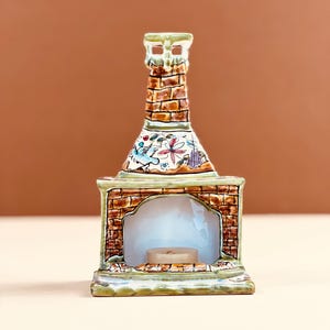 May include: A ceramic candle holder shaped like a fireplace with a chimney. The fireplace is made of brown bricks and has a decorative floral and bird design. A lit candle sits inside the fireplace.