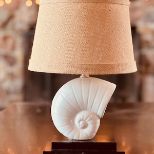 May include: A decorative table lamp featuring a white seashell base and a beige burlap lampshade. The lamp has a dark brown rectangular base and a silver-colored metal fitting. The lamp is designed for indoor use.