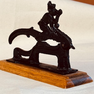 May include: A black cast iron sculpture of a person riding a mythical creature, mounted on a rectangular wooden base. The figure is detailed, with the person holding something to their mouth. The base is a light brown color.