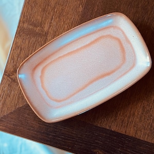May include: A rectangular ceramic serving dish with a pearlescent finish. The dish features a gradient of colors, transitioning from a light peach center to a white and light blue edge. The dish is placed on a dark wooden surface.
