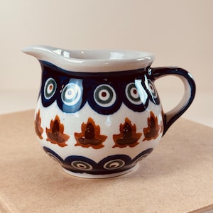 May include: A small, white ceramic creamer with a blue and brown floral pattern. The creamer has a rounded body and a small handle.