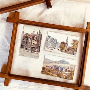 May include: A rectangular wooden floating frame with a dark brown finish containing three vintage postcards. The postcards feature detailed illustrations of historic city streetscapes and a large stone building set against a mountainous landscape.