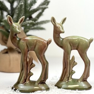 May include: Two ceramic deer figurines in shades of green and brown. The deer have a stylized design with slender legs and a curved base. The figurines are positioned on a white surface, with a blurred Christmas tree in the background.