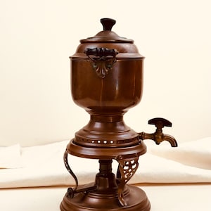 May include: A copper samovar featuring a rounded body, a lid with a dark knob, and side handles. The base includes a decorative metal stand with cut-out details and a brass spigot with a handle extending from the side for dispensing liquid.