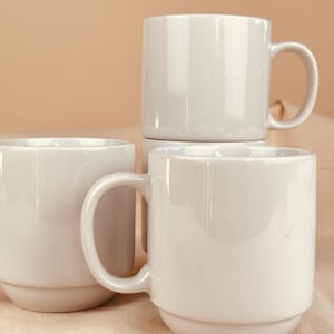 May include: Three stacked, off-white ceramic mugs with rounded handles. The mugs have a simple, minimalist design and a glossy finish. The mugs are arranged on a neutral-colored surface, creating a clean and modern aesthetic.
